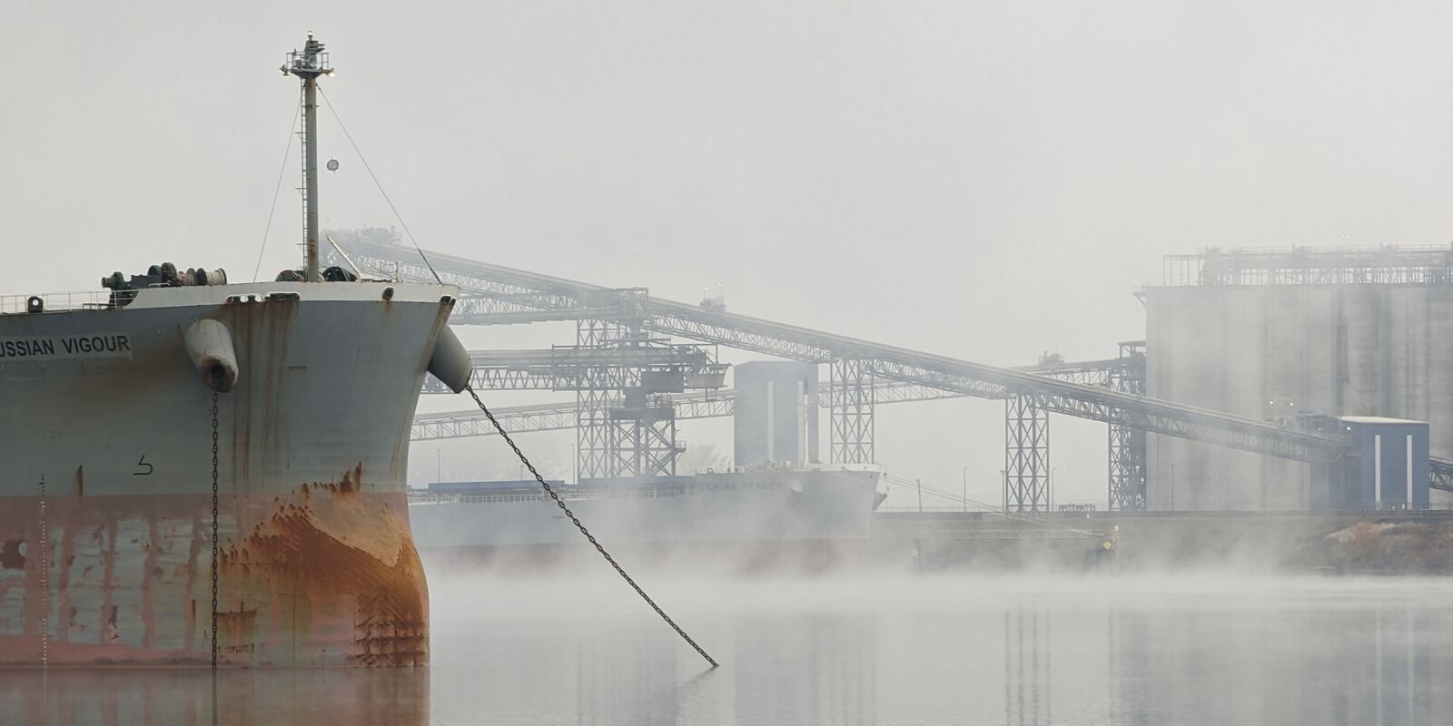 The bow of a ship moored at a grain terminal in morning fog.